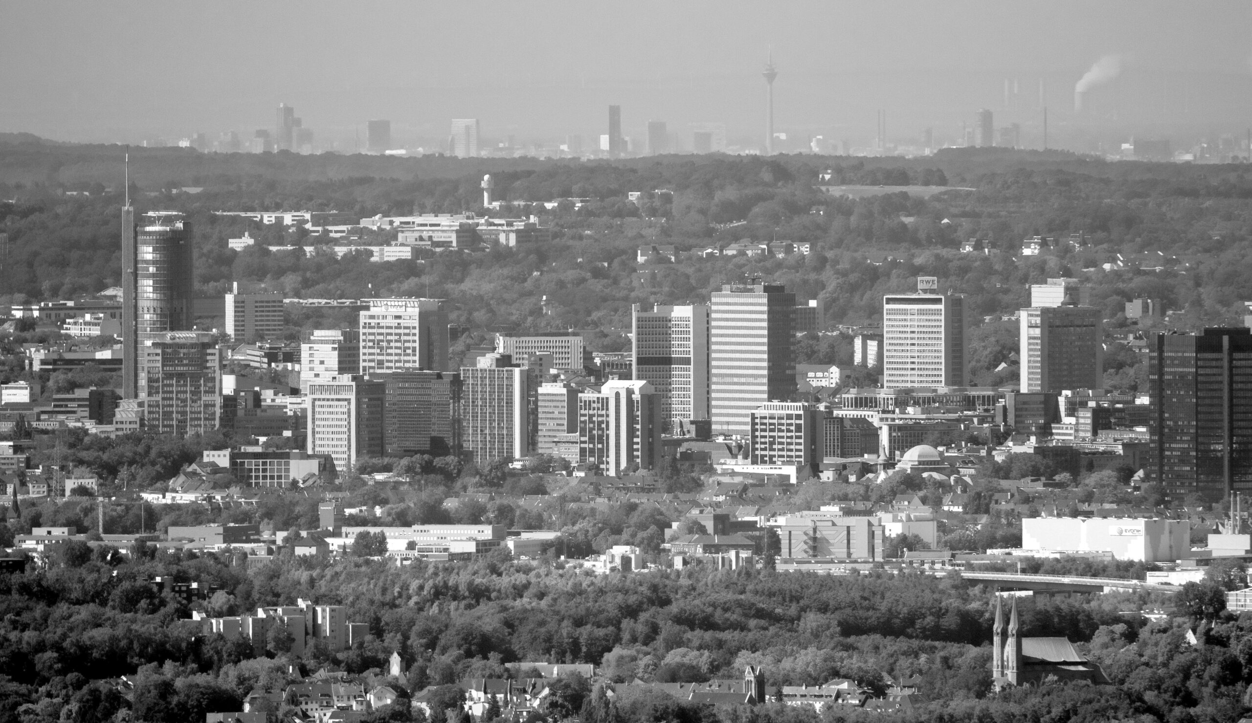 Schauen Sie mal ganz genau hin. Auf diesem Luftbild hat Hans Blossey bei außergewöhnlich klarer Sicht mit einem Teleobjektiv die Skyline von gleich zwei Städten eingefangen: Vorne im Bild sind das Essener Hochhausviertel mit RWE-Turm (links), neuer Schenker-Zentrale und Rheinstahl-Haus (rechts) sowie das Essener Rathaus (vorne, rechts außen) zu sehen. Am Horizont grüßt obendrein der Düsseldorfer Rheinturm. Der Landtag direkt daneben ist zwar nicht in Sicht, aber das Dreischeibenhaus (ehemaligen Thyssen-Krupp-Zentrale, etwas weiter links) ist ebenso deutlich zu erkennen wie das Stadttor (Sitz der Staatskanzlei) und der Victoria-Tower (Sitz der Ergo-Versicherung). Rechts im Bild ist oben neben einer Rauchwolke sogar eine Rheinquerung zu sehen: die Theodor-Heuss-Brücke. Das Flugzeug mit Hans Blossey war in diesem Moment übrigens über Gelsenkirchen (südlich des Zoom-Zoos) unterwegs. Luftlinie von dort bis zum Essener Rathaus: etwa 11,5 Kilometer. Die direkte Distanz bis nach Düsseldorf: etwa 40 Kilometer. Noch zwei Orientierungspunkte im Essener Bildvordergrund: Hinter dem Grüngürtel ragt halblinks der Turm der Feuerwehrhauptwache an der Eisernen Hand empor, vorne rechts im Bild der Doppelturm der Kirche St. Nikolaus in Stoppenberg.