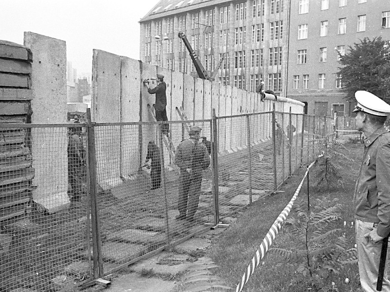 Direkt an der Sektorenlinie bewachten Grenzsoldaten mit mobilem Sicherungszaun die Mauerarbeiten. Am linken Rand steht noch die ältere Betonschichtmauer aus horizontalen massiven Wegeplatten. Auf West-Berliner Seite beobachtete ein US-Militärpolizist das Geschehen.