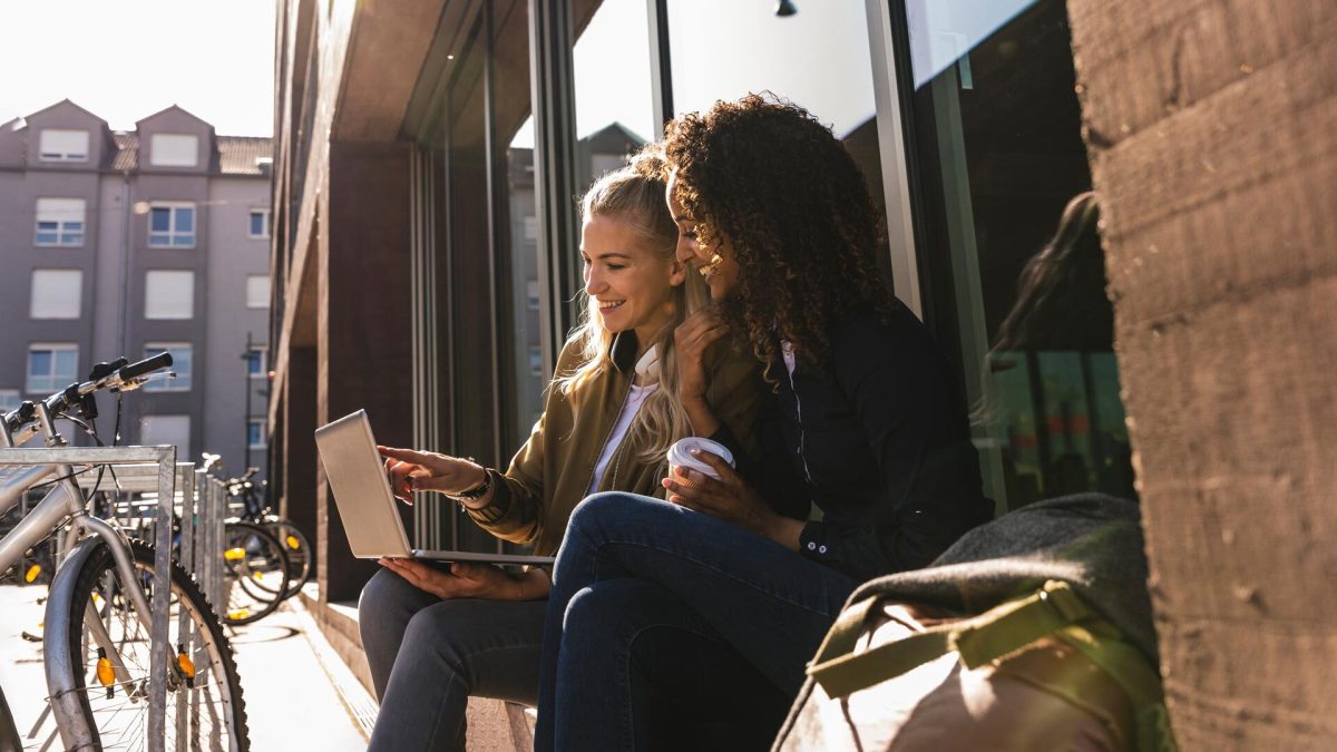 Zwei junge Frauen sitzen vor einer Universitätsbibliothek vor dem Laptop