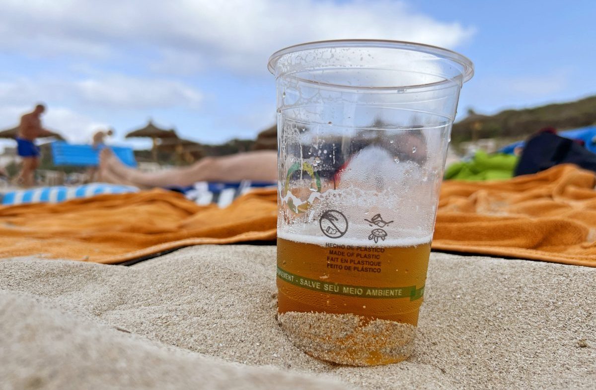 halbgefÃ¼llter Bierbecher am Strand von Mallorca, Urlauber auf HandtÃ¼chern im Hintergrund