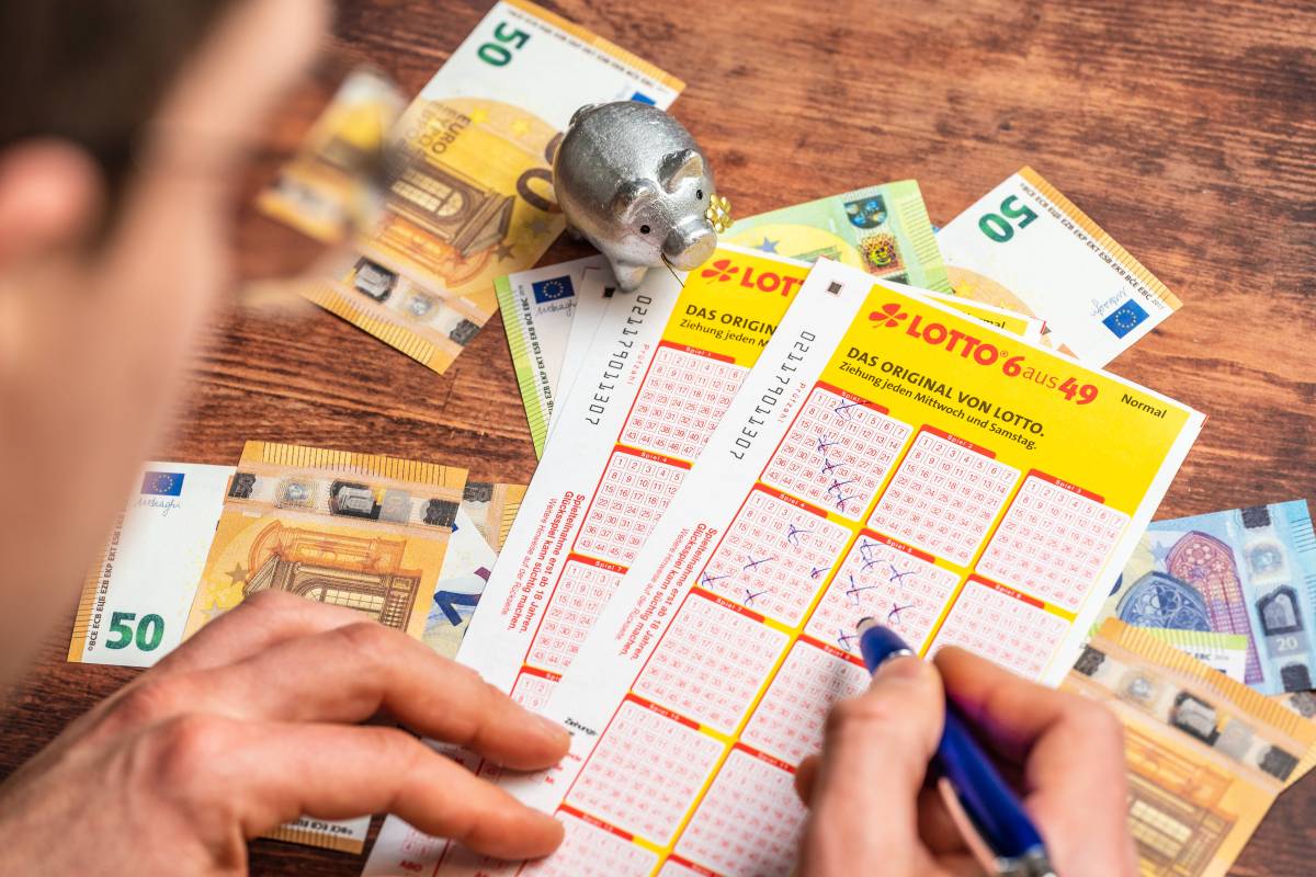 Ein Mann füllt einen Lottoschein aus, um den herum mehrere Euro-Geldscheine und ein Glücksschwein liegen.
