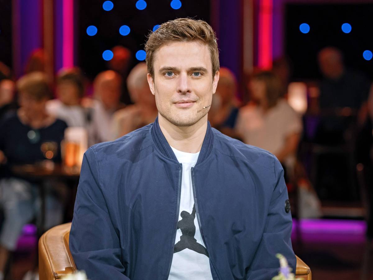 Auf dem Bild ist Tagesschau-Moderator und Schriftsteller Constantin Schreiber. Er sitzt in einem Fernsehstudio. Er trägt ein weißes T-Shirt und darüber eine blaue Bomberjacke. Er hat braune hochgegelte Haare und an seiner Wange ein Mikrofon angebracht.