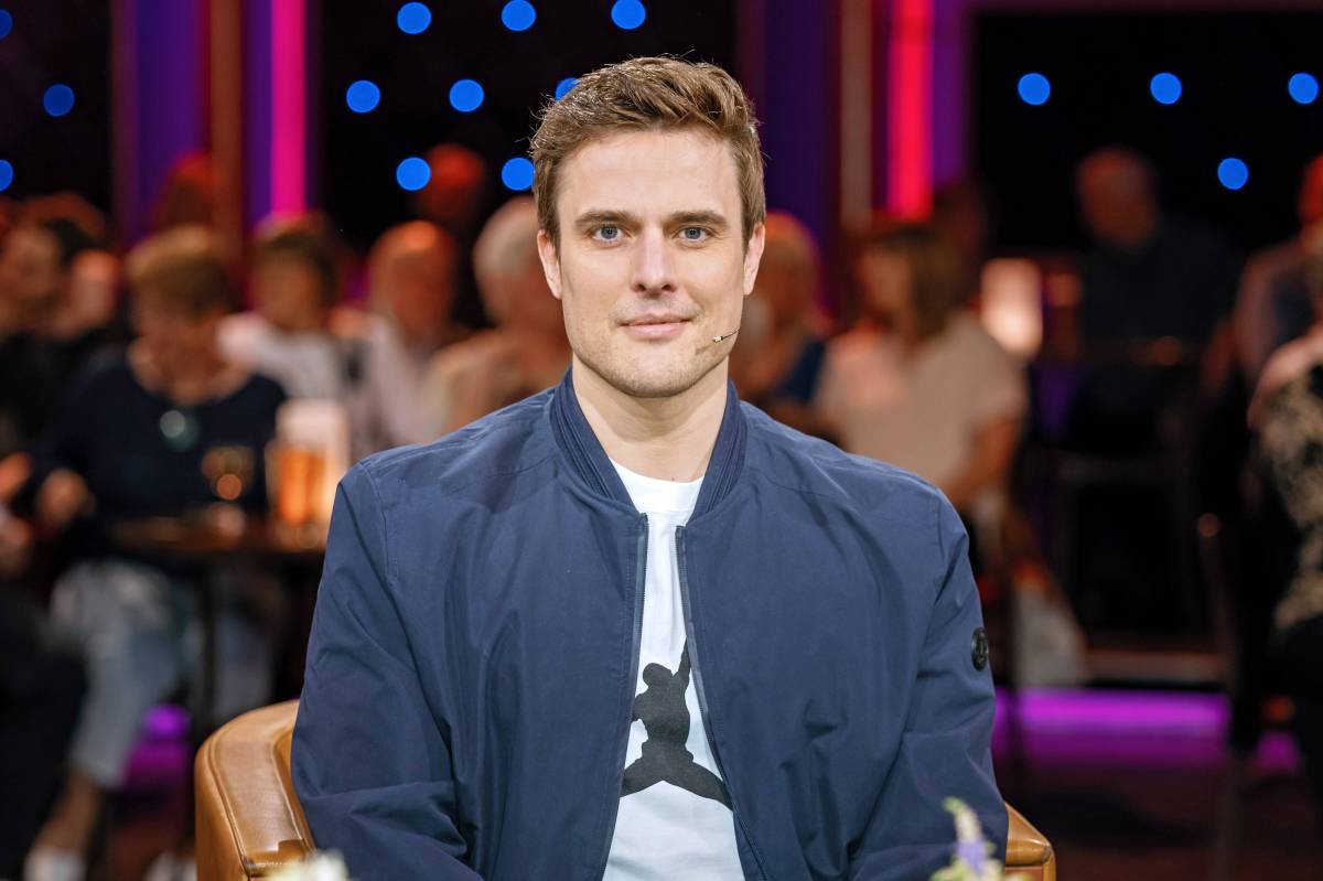 Auf dem Bild ist Tagesschau-Moderator und Schriftsteller Constantin Schreiber. Er sitzt in einem Fernsehstudio. Er trägt ein weißes T-Shirt und darüber eine blaue Bomberjacke. Er hat braune hochgegelte Haare und an seiner Wange ein Mikrofon angebracht.