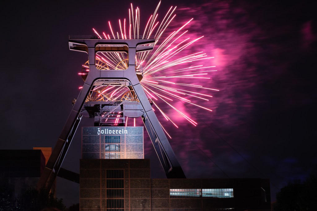 Ein Turm der Zeche Zollverein in Essen hinter dem ein Feuerwerk erstrahlt. Die Zeche Zollverein in Essen ist eine der bekanntesten Sehenswürdigkeiten der Stadt.